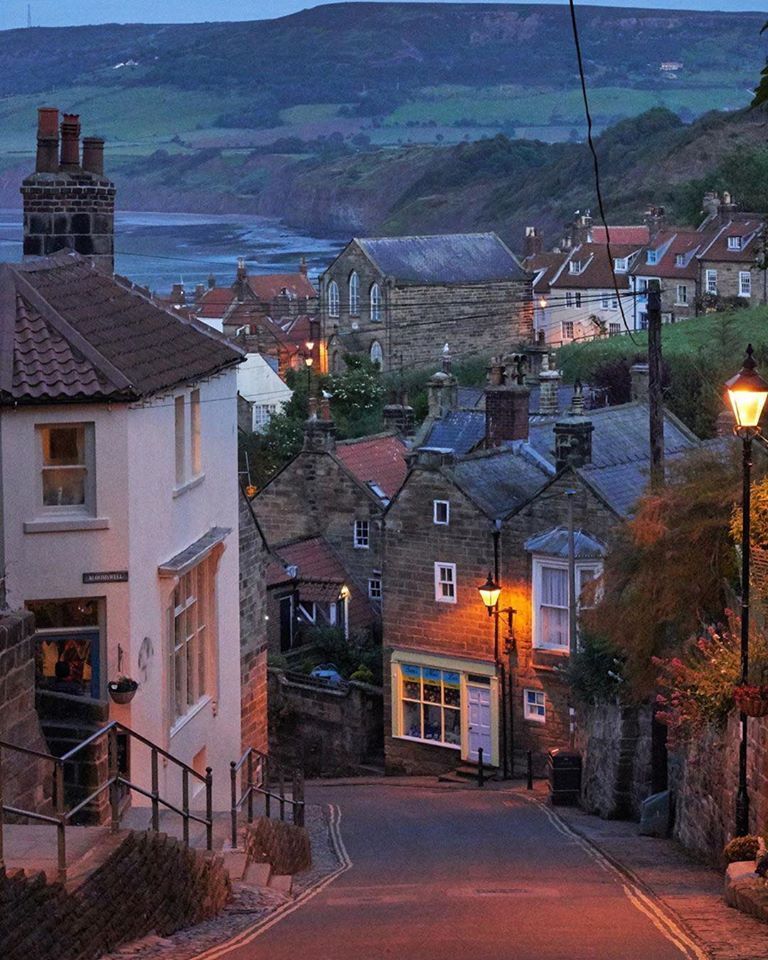 Narrow lane through a stone-built English coastal village at dusk with glowing street lamps and sea in background