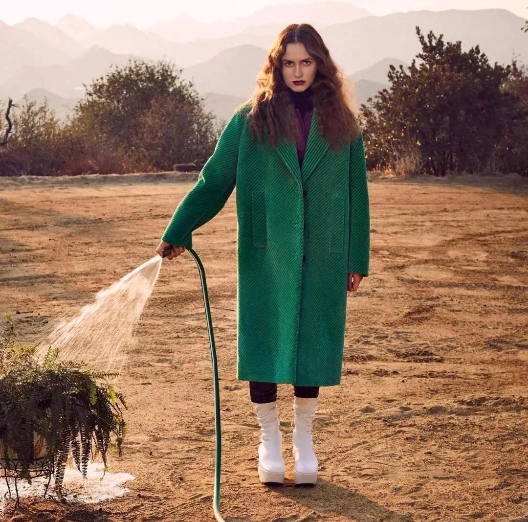 Woman wearing long green coat standing in open field with mountain backdrop