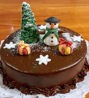 Chocolate cake decorated with a snowman, evergreen tree, gift boxes, and powdered sugar snowflakes