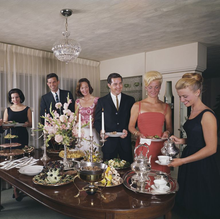 Six people in formal 1960s attire at an elegant cocktail party around a decorated table with chandelier overhead