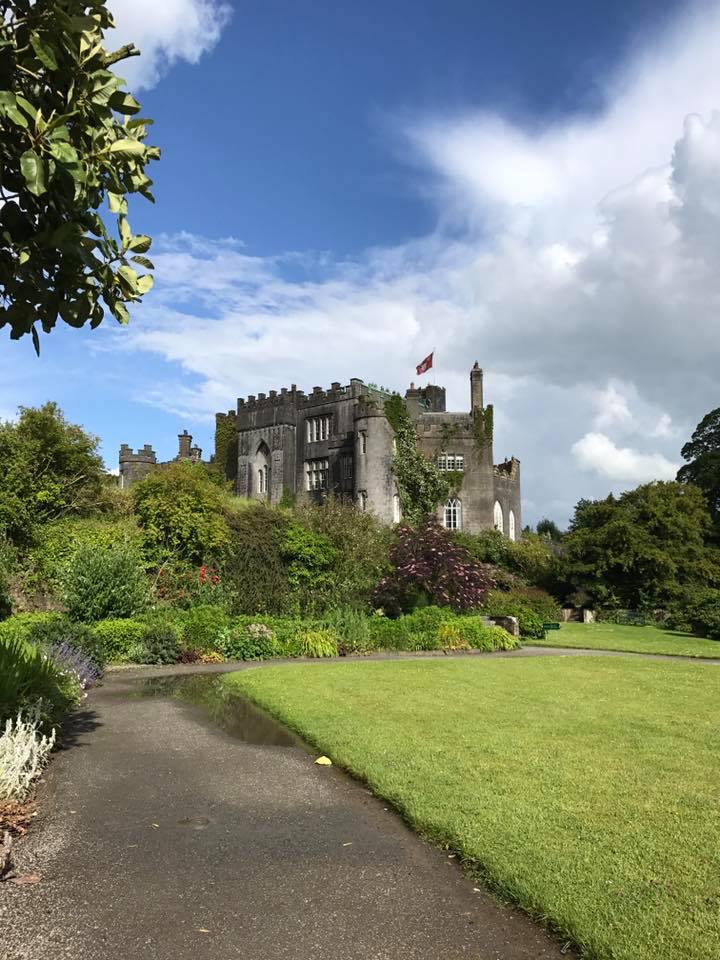 Historic stone castle with tower and flag surrounded by green gardens and lawn under blue sky with clouds