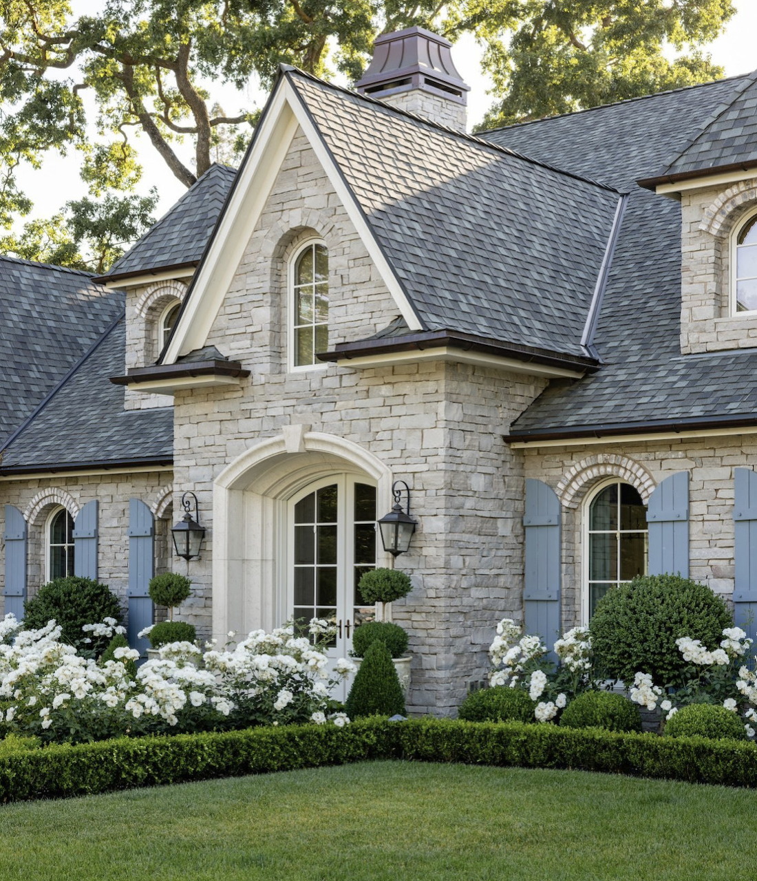 Stone cottage exterior with white hydrangeas, manicured lawn, and gray slate roof under tree canopy