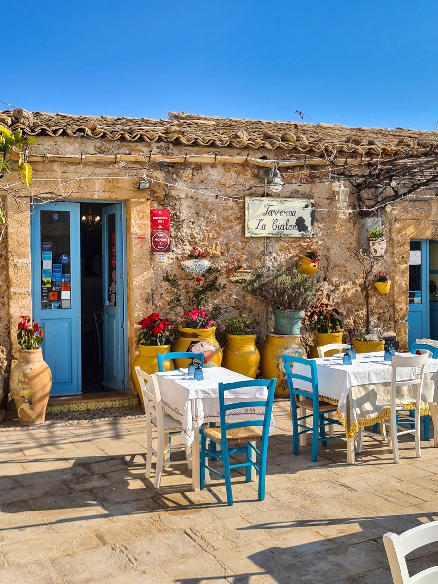Rustic taverna terrace with colorful chairs, yellow pots, and blue door against weathered stone wall