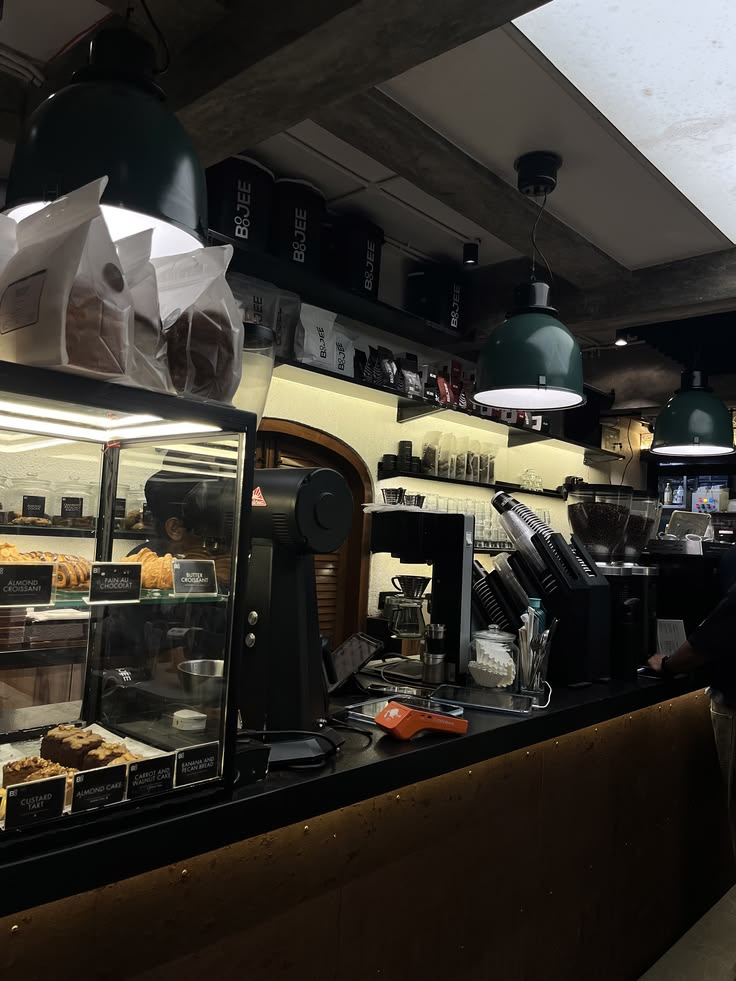 Dark café interior with espresso machine, pastry display case, and green pendant lights above the counter