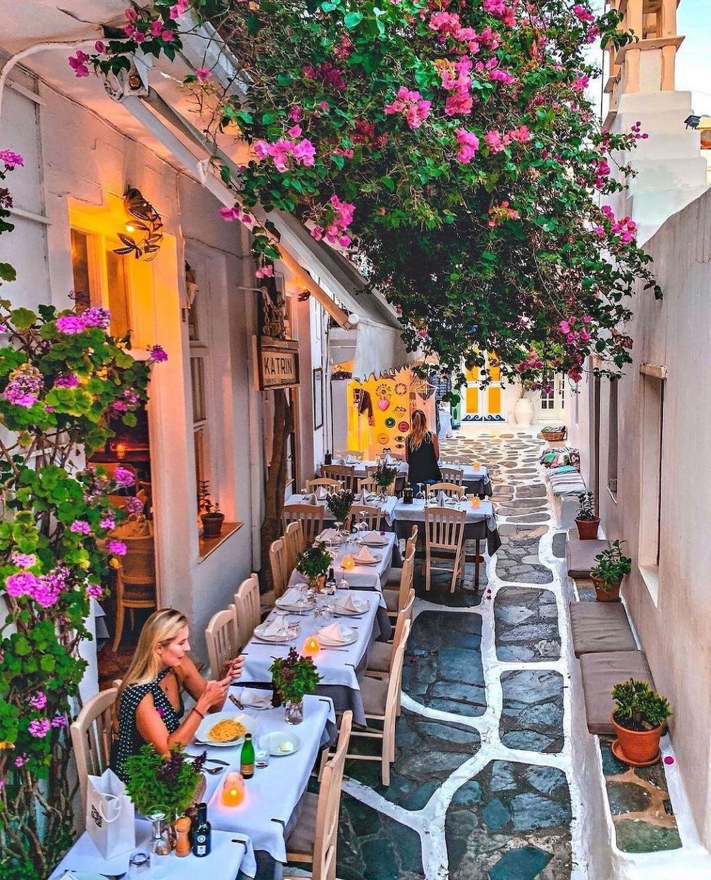 Picturesque Greek alley with pink flowers, white buildings, and outdoor cafe tables on cobblestone street