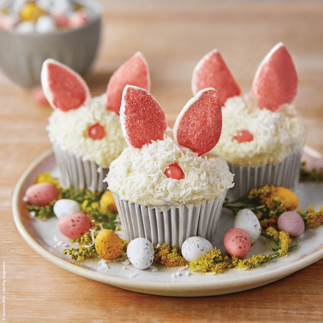 Four white frosted cupcakes decorated as Easter bunnies with pink candy ears and candy eggs arranged on a plate