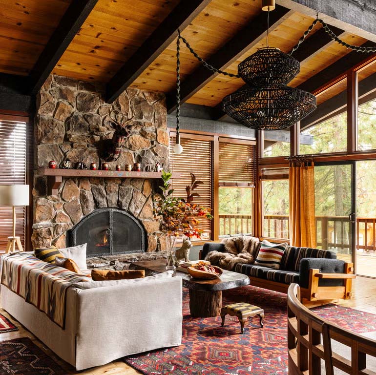 Rustic cabin living room with stone fireplace, wooden ceiling beams, and large windows overlooking trees