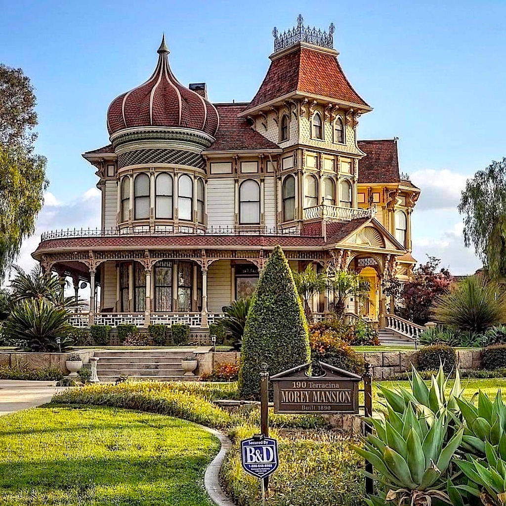 Grand Victorian mansion with red roof, turrets, ornate trim, and wraparound porch surrounded by landscaped garden