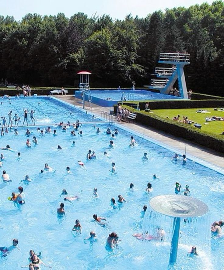 Aerial view of crowded outdoor pool with swimmers, diving platforms, and water slides surrounded by trees