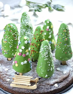 Green Christmas tree cakes with sprinkles and powdered sugar on a round board beside a tiny sled