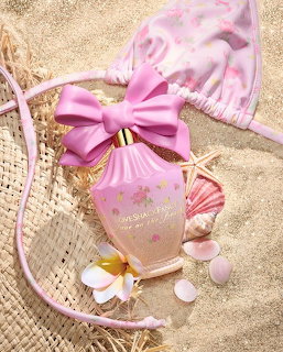 Pink perfume bottle on sand with bikini top, straw hat, flower, and seashells