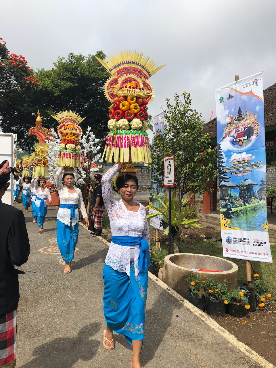 Women in blue and white traditional dress carrying tall colorful flower offerings during Balinese festival procession
