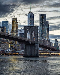 Brooklyn Bridge with Manhattan skyscrapers and One World Trade Center against a dramatic cloudy sky at dusk