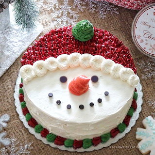 Round white cake decorated to look like a snowman with candy face, red beads, green hat topper, and holiday decor