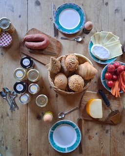 Overhead breakfast table with bread rolls, strawberries, cheese, jam jars, eggs, and plates