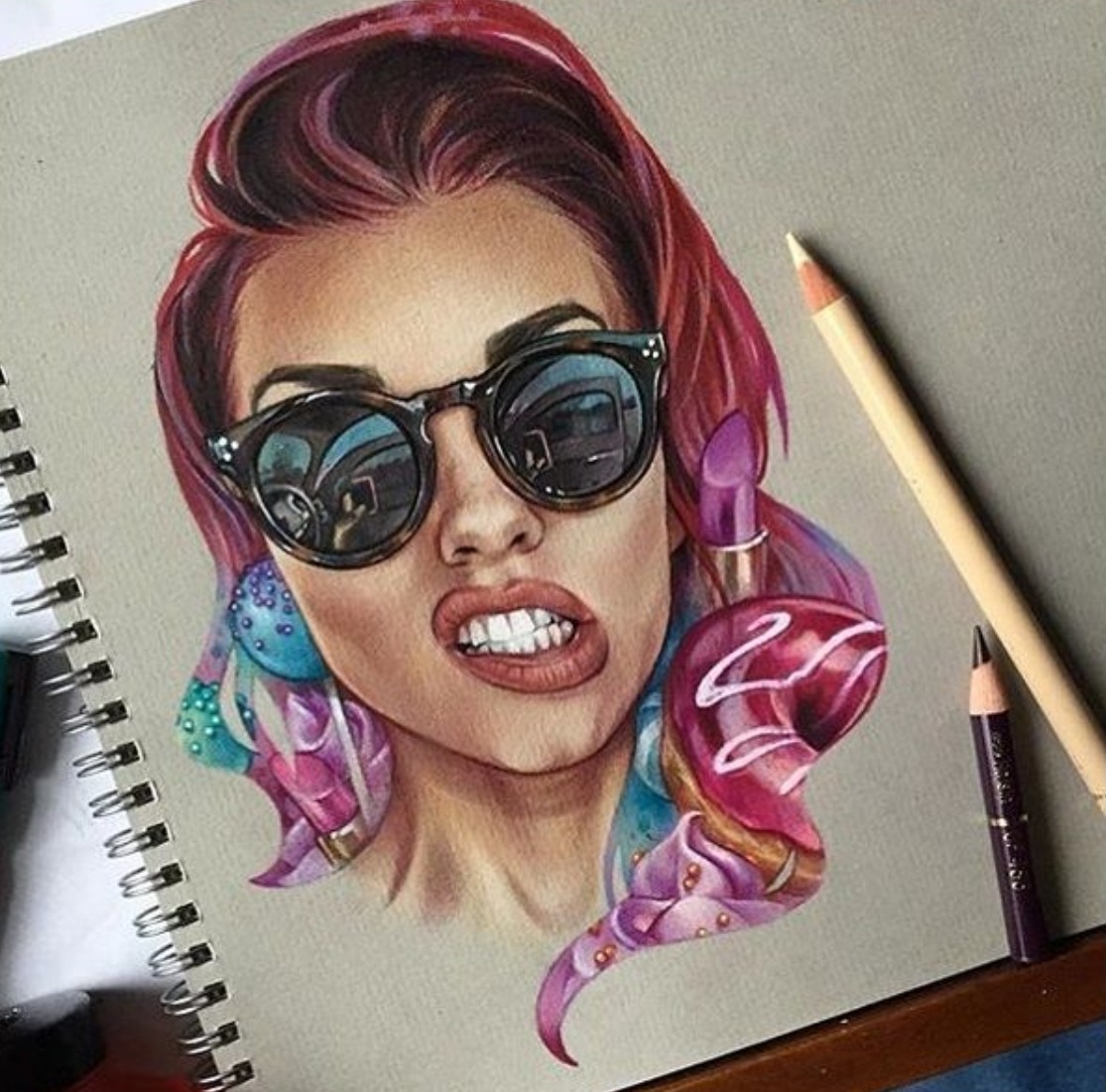 Colorful portrait drawing of a woman wearing sunglasses on a sketchbook page with pencils nearby