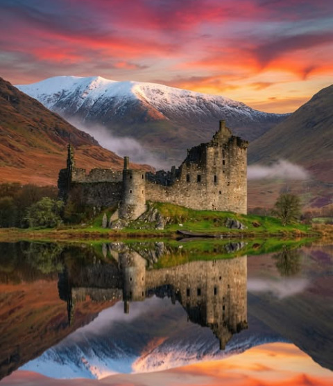 Ruined stone castle by a lake with mountain backdrop and vivid sunset reflection