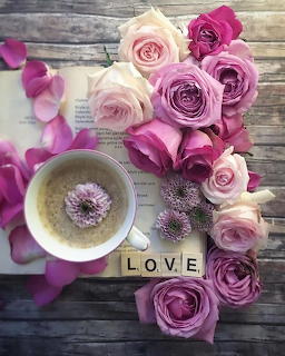 Pink and purple roses arranged on a wooden table with a coffee cup, open book, petals, and LOVE Scrabble tiles