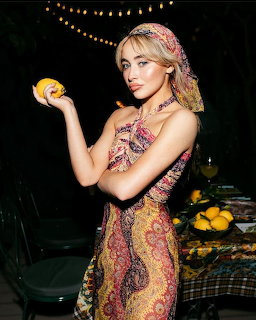 Blonde woman in paisley dress and headscarf holding a lemon at a nightime outdoor setting