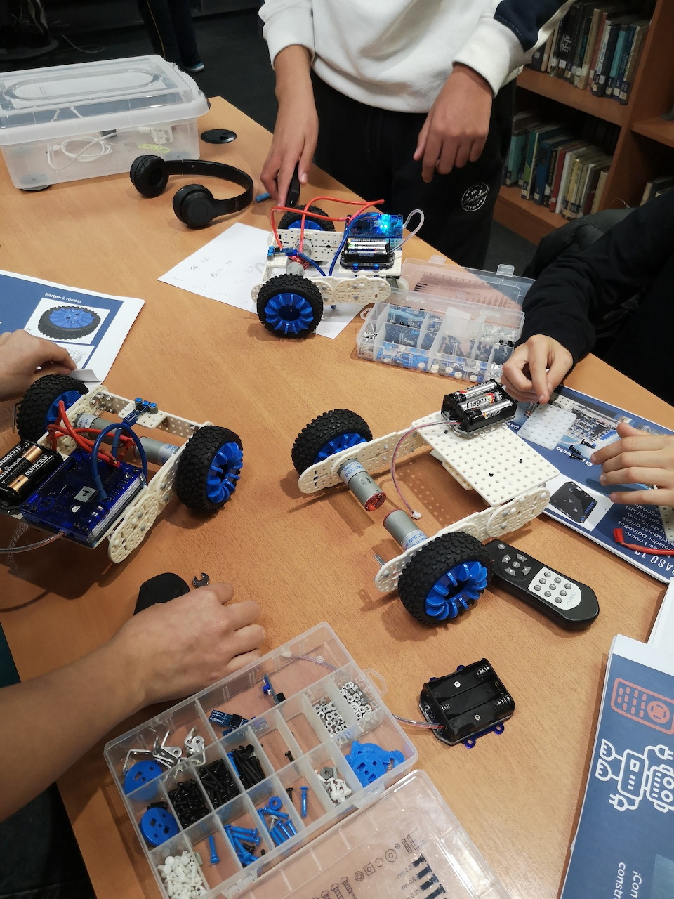Students building wheeled robots at a table with electronic parts and instruction booklets in a classroom.