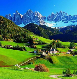 Alpine village with green meadows, church, and snow-capped mountains under a blue sky