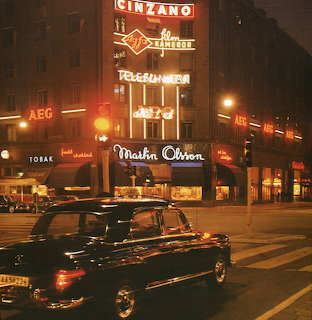 Vintage city street at night with a classic black car, neon signs, and illuminated shops