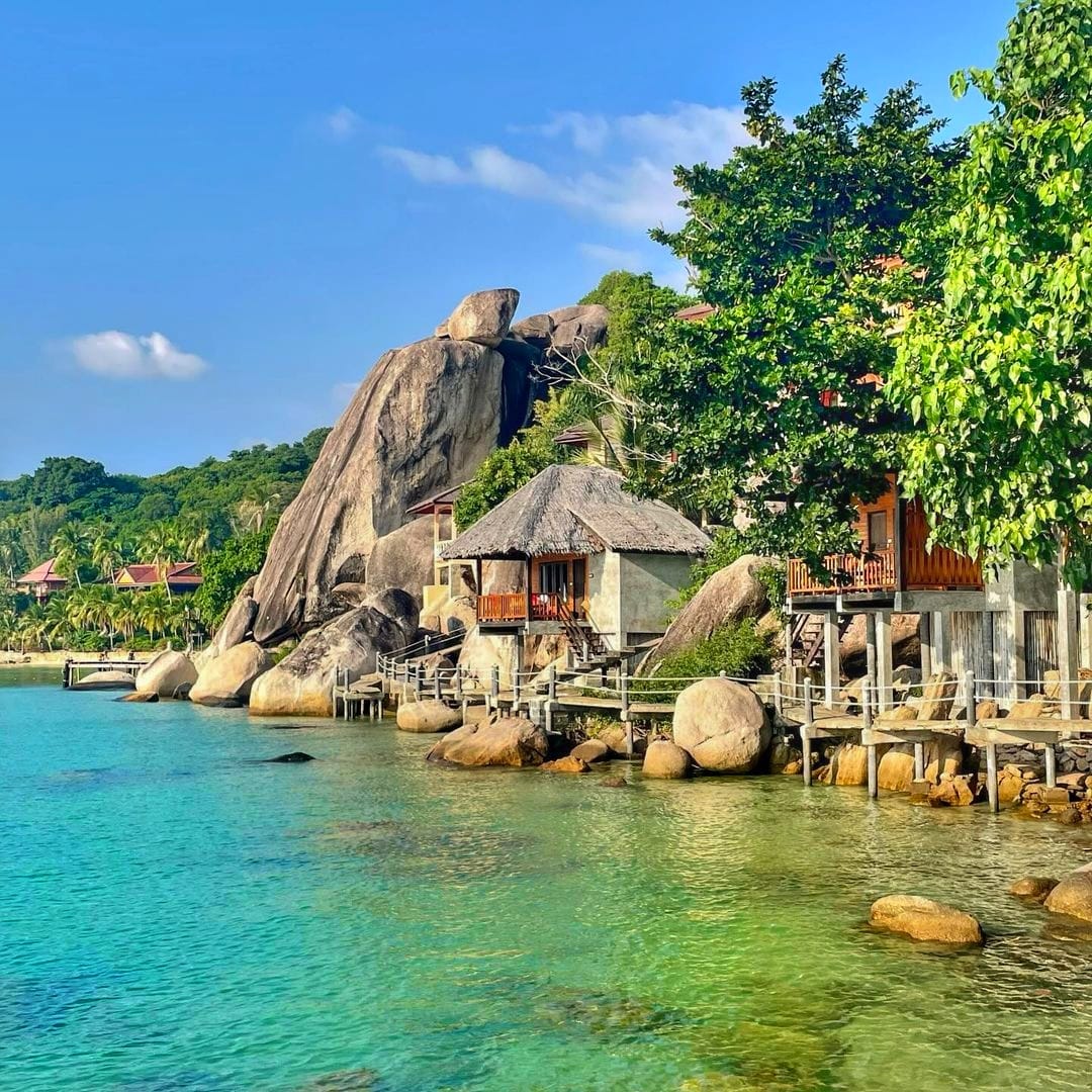 Wooden huts by granite boulders on a clear turquoise shore in Koh Tao, Thailand