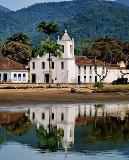 White colonial church with bell tower reflecting in tranquil waterfront, lush mountains behind