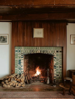 Lit brick fireplace with green tiles in a wood-paneled living room with stacked firewood