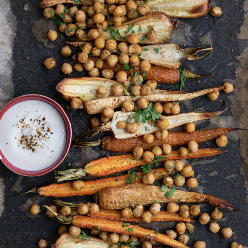 Overhead view of roasted carrots, endives, chickpeas, and a bowl of creamy sauce on a dark tray