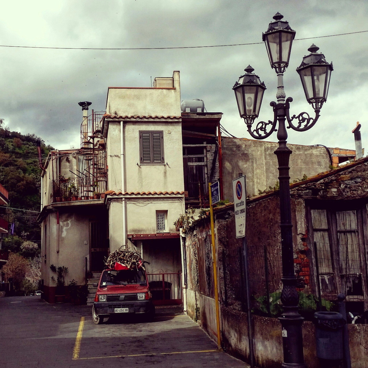 Narrow old street with worn buildings, a parked pickup truck, and tall street lamps under cloudy skies
