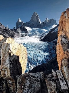 Jagged mountain peaks above a blue glacier framed by rocky cliffs and a clear sky