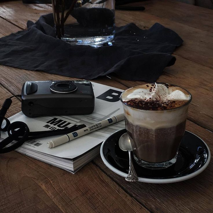 Glass of foamy coffee on a saucer beside pens, a notebook, and a camera on a wooden table