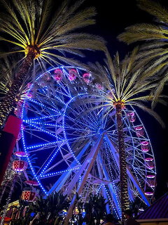 Large ferris wheel lit with vibrant blue and purple lights at night, flanked by silhouetted palm trees
