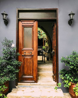 Open rustic wooden door with arched garden view, flanked by poted plants and wall lanterns