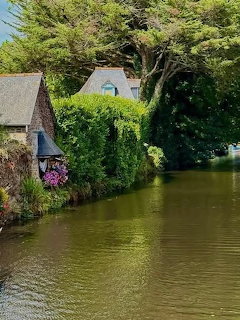 Stone cottage beside a calm green river with lush trees and a flower-decorated boat at the waterside