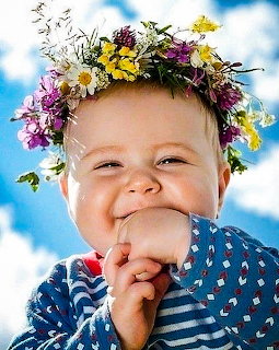 Happy toddler with colorful flower wreath crown smiling outdoors against blue sky