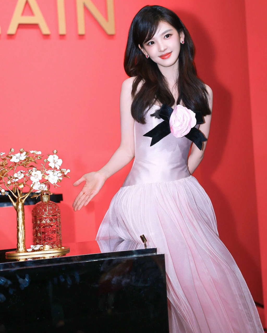Woman in a pale pink dress posing against a red backdrop beside flowers and a table