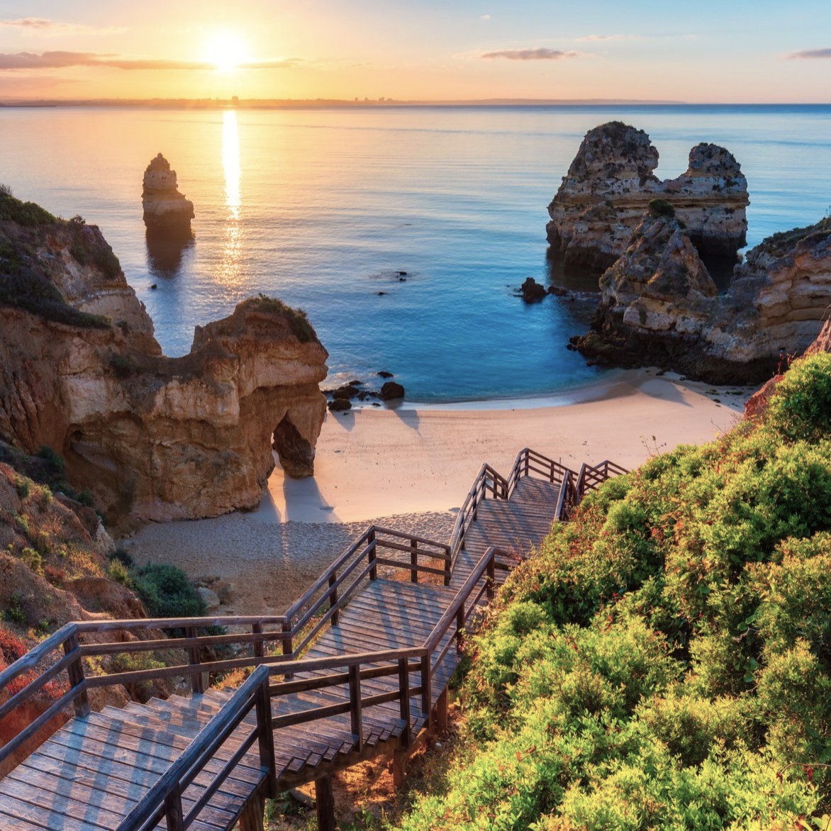 Wooden stairs leading to secluded beach with dramatic rock formations and sunset over calm ocean in Algarve, Portugal