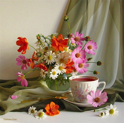 Still life with pink, orange, and white wildflowers in a glass vase next to a porcelain tea cup on green draped fabric