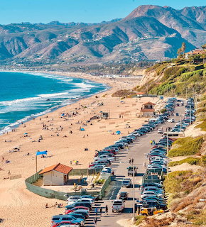 Coastal highway with parked cars alongside a busy sandy beach with mountains in the background