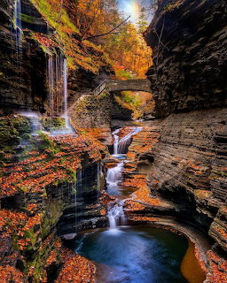 Waterfall cascading through layered rock gorge with autumn leaves and stone bridge above