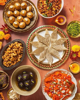 Flat lay of assorted Indian sweets and snacks on a teracotta-colored surface with flowers and diyas
