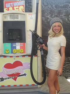 Smiling woman in a white dress standing beside a vintage gas pump holding the nozzle