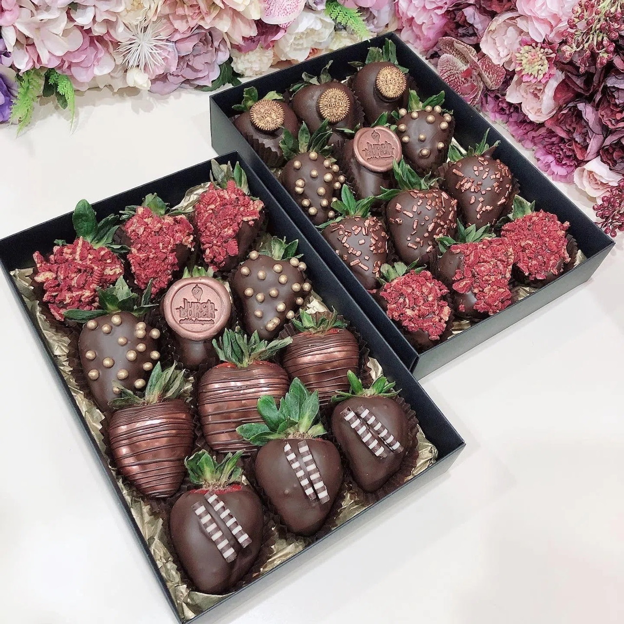 Chocolate-covered strawberries decorated with white drizzles and pearls in black gift boxes surrounded by pink flowers