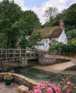 White thatched cottage with stone bridge over water, pink flowers in foreground, trees surrounding the building
