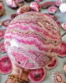 Polished pink rhodochrosite sphere with wavy banding patterns held in hand over rhodochrosite slices
