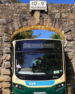 Green and gold double-decker bus passing under a stone arch with headroom warning sign