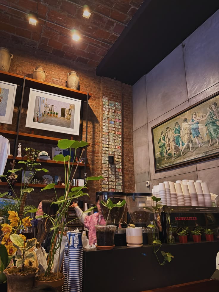 Cafe interior with brick walls, lush potted plants, framed paintings, and a counter with stacked cups