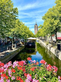 Canal in Delft with vibrant flower boxes, moored boats, trees, and a historic church tower in the background